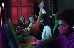 Four young people sit at a row of computers in a gaming arena. Two in the middle, wearing headsets, smile and high-five each other while the others focus on their screens, surrounded by colorful lights.