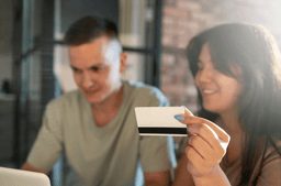A woman holds a credit card while sitting next to a man. Both are looking at a laptop screen, suggesting they are making an online purchase or payment together. The setting appears to be indoors with natural light.