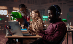 A man wearing headphones works on a laptop at a desk in a library, holding a pen. In the background, two women collaborate, one holding a blue folder. The room is softly lit with green desk lamps.