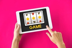 Hands holding a tablet displaying a slot machine game with three reels showing stars and the number 7, against a bright pink background.