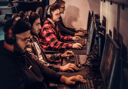 Four people wearing headphones sit side by side at desks, focused on computer screens, using keyboards and mice, in a dimly lit room, likely participating in a gaming or esports event.