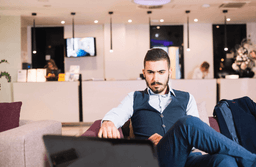 A man in business attire sits on a couch in a modern lobby, focused on a laptop in front of him. Reception desk and staff are visible in the background under hanging lights.