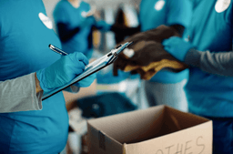 Volunteers in blue shirts and gloves sort clothes and write on a clipboard near a box labeled "clothes" in a donation center.