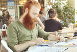 A man with a long red beard and green shirt sits at a table in a café, looking at documents and holding a pencil. Papers and a glass of coffee are on the table; other people are seated in the background.