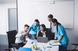A group of medical professionals and a businesswoman sit and stand around a table, reviewing X-rays and documents in a modern office with a laptop, fruit bowl, and whiteboard visible.