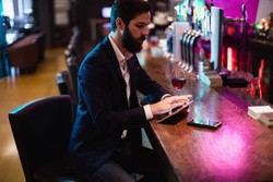 A man with a beard sits at a bar counter using a tablet. There is a glass of red wine and a smartphone on the wooden counter beside him. The bar background is dimly lit with colorful lights.