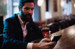 A man with a beard, wearing a suit, sits at a bar holding a glass of red wine in one hand and looking at a smartphone in the other. The background is softly lit and out of focus.